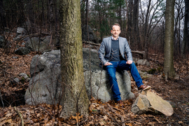 Fournier sits on a large grey rock in the woods. The textures of the trees, rocks, and dead leaves contrasts with Fournier’s blue jeans.