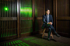 In Song Kim is seated in chair in a room with wooden slatted walls. Through the slats, a green light shines and casts interesting shadows on the ground.