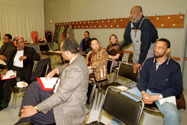 About nine people sit in a classroom. Mel King is standing near the back speaking, and people are turned to listen to him.