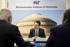 President Yoon Suk Yeol sits at a table in front of a microphone, speaking. Two people are facing him, with their backs to the camera, and behind him is a screen showing a photo of MIT’s Great Dome, with the MIT logo and the words “Massachusetts Institute of Technology.” 