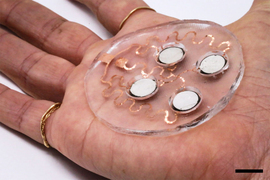 On the palm of a person’s hand is a unique circular device. The base looks like clear gel pad with 4 circular white sensors on it. Wavey copper lines attach the sensors to each other.