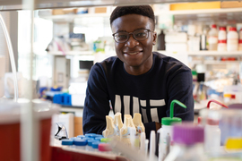 Victor Damptey wears a blue MIT sweater, and smiles in his lab full of items. The photo is taken across the table divider, so he is framed by a shelf, top, and the table. Models of bones, and many bottles and canisters are on the table.