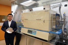 Jiadi Zhu wears gloves while holding an octagon-shaped plastic case containing 8-inch wafer. On right, a furnace resembling a deep freezer sits in glass casing.
