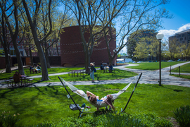 The grassy area outside of the Chapel at MIT shows several people working at tables and benches, along with paved paths. A person walks along a path while two people lay in a hammock under the sun.