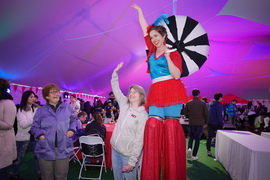 Under a large tent, Sally Kornbluth, casually dressed, smiles and waves while standing next to a performer on stilts wearing bright red and blue clothing.