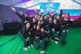 Student dancers wearing black tops and green pants pose together in a large group, smiling with arms outstretched.