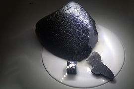 The meteorite is dark black and shiny, with a rough texture. A chip of the meteorite is next to it, and a cube says “1 CM” on it.