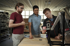 Elijah Stanger-Jones, Hongmin Kim, and Andrew SaLoutos look at a metal robot gripper that resembles an excavator. The gripper holds a pink cup as it sits atop a wooden table.