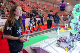 Inside the Johnson Athletics Center, Sarah Stoops wears a 2.007 shirt as she uses a remote control to control a metal robot with 4 yellow wheels and lots of wires on top. In the background are balloons, a referee in a striped shirt, and people watching in the stands.