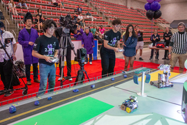 Ethan Chang and Nikolaos Tsakiris stand side by side, using remote controls to control their robots on the game board. Wearing purple lab coats, professors Amos Winter and Sangbae Kim look on, as do others in the crowd.