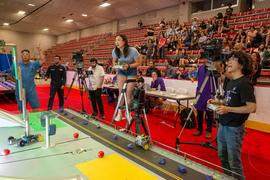 Alexandra Lee and Ethan Chang excitedly watch their robots. Lee is on a ladder with a remote control.  Behind them, the crowd in the bleachers looks on with smiles and intensity.