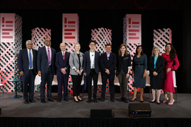 Participants in the panel discussion stand side-by-side on the stage, posing for group photo. Square columns with the MIT logo rise up behind them.
