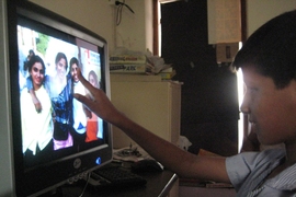 A child is seated in front of a computer screen that has a photo of 4 people smiling. The child’s finger points directly at a person’s face.