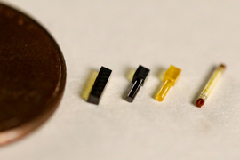 Next to a penny are 4 very tiny devices that are a similar shape to one another. One is rectangular, 2 are rod-like with a cube at one end, and the last is a rod with red ends.