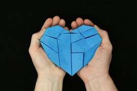 Two hands hold a kirigami blue heart above a black background
