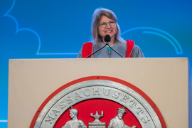 President Kornbluth, wearing grey and red academic regalia, speaks at a podium with a large MIT seal on the front.
