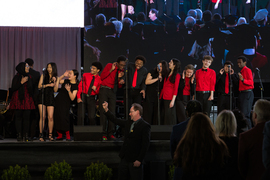 15 singers wear red and black. They are comically reacting to a loud noise made by a singer on the left, laughing while covering their ears. 