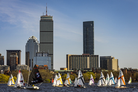 About 20 colorful MIT sailboats on the water, with the Boston skyline in background.