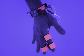 A hand shows the index and middle fingers wrapped in tape and connected to an electric wristband through a set of wires.