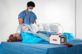A doctor works on a patient using the SurgiBox. The Surgibox is made of translucent material that covers the patient’s body, with holes for the doctor’s gloved hands. A white and red box is next to the patient and inflates the bubble.