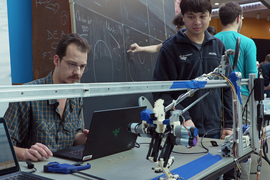 Students at a table, with a laptop and their chicken robot. They watch the robot walk on a blue line.