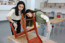 2 students paint a wooden chair inside a classroom.