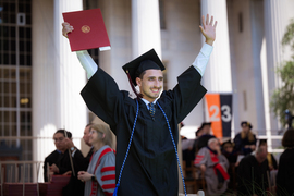 A male graduate wearing cap and gown, smiling and holding his diploma in the air.