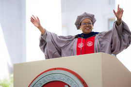 Chancellor Melissa Nobles in academic regalia speaking at a podium with her arms raised.