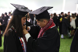 Two graduates in black robes helping each other get ready.