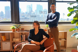 Shara Ticku, seated, and David Heller pose for a portrait. A window showing the Cambridge cityscape and two cube storage shelves are behind them.