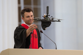 Mark Rober has attached his graduation cap to a flying drone, and he gestures to it as it flies away.