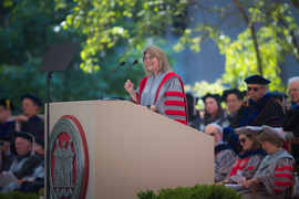 Sally Kornbluth, wearing academic regalia, speaks at a podium with a red and silver MIT seal on the front, with green foliage and seated faculty in background.