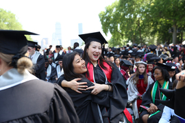 Two graduates in caps and gowns hug and smile while surrounded by other graduates. Trees and Boston skyline are in the background.