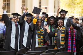 Seven graduates in caps and gowns, with some wearing kente stoles, smile at the camera with arms raised. The columns of Killian Court are in background.