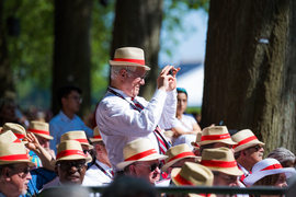 Wearing a straw hat with a red stripe, an alumnus stands up and takes a photo while surrounded by seated alumni wearing the same hats.