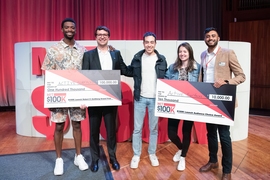 5 people stand on stage holding 2 gigantic checks that say they’ve won $100k and $10k. In the background, a large red and beige sculpture says MIT $100K.
