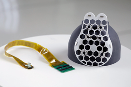 On the left of the photo, a yellow strip-cable has a green circuit board on one end, and a small ultrasound tracker on the other. On the right, the white device is teardrop shaped and has a hexagonal grid of holes where the tracker can be placed.