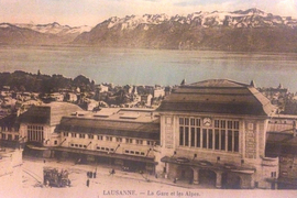 A pink-hued faded vintage postcard shows the buildings of Lausanne and the Alps in background, and says “LAUSANNE. La Gare et les Alpes.”