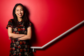 Lisa Ho smiles while leaning against a red stairwell wall. A grey handrail diagonally goes up.