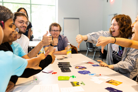 About 8 people at a table smile and give a “thumbs-up” and “thumbs-down” while playing a game with black cards.