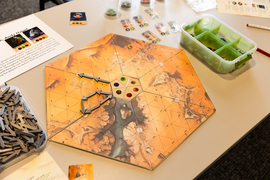 A hexagon-shaped game looks like an orange planet’s surface. Game pieces include grey arrows little card with illustrations on them.