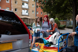 A group of 3 look at trifold brochure while two carts of clothes, luggage, bags, a van, and an apartment building surround them. A family member is at the right with a rolling luggage.