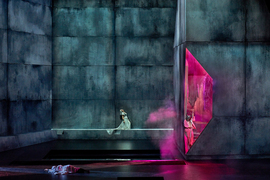 3 actors are dwarfed by the gigantic grey and black stage. A person lies on the ground, and a woman looks up dramatically. Pink light shines on a person wearing a pink suit and a demonic mask.
