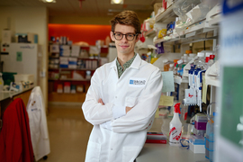 Tzouanas wears a Broad Institute lab coat and stands with arms crossed, leaning against a countertop inside a lab. Lots containers, boxes, and lab equipment fill the shelves around the lab.