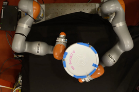Top view of a demo showing 2 robot arms, manipulating a white bucket labeled “Mr. Bucket.”