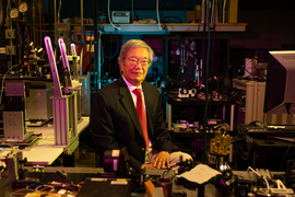 James Fujimoto sits in a crowded laboratory surrounded by imaging equipment. The lab is darkly lit with low fuschia, gold, and teal lights. 