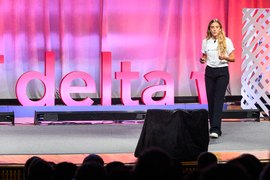 A woman wears a “Medikana” shirt while presenting on stage. The stage has a red sculpture that says, “MIT delta v.”
