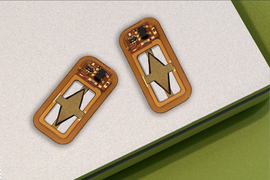 Two tiny rectangular devices have curved edges. The devices are orange-yellow and are made of a circuit board and soldered pieces, including a diamond-shaped piece of material in the middle.