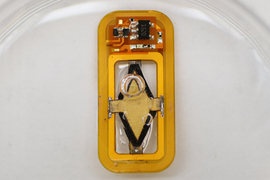 A tiny rectangular device has curved edges and is in water, and bubbles form on top. The device is orange-yellow and has a circuit board and soldered pieces, including a diamond-shaped piece of material in the middle.