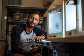 Faraz Faruqi holds a 3D-printed figurine and sits next to two brightly lit 3D-printers inside the lab.